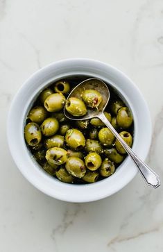 a white bowl filled with green olives on top of a marble counter next to a spoon