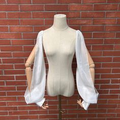 a mannequin wearing a white top with open sleeves on a wooden stand in front of a brick wall