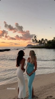 two women standing on top of a sandy beach next to the ocean at sunset with palm trees in the background