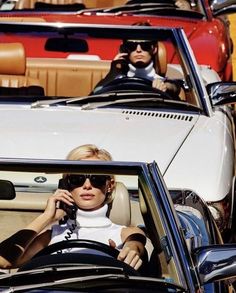 two people sitting in a convertible car talking on the phone