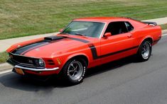 an orange muscle car with black stripes on the hood driving down a street next to grass