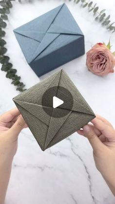 someone holding an origami envelope in front of some flowers and a blue box