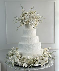 a wedding cake with white flowers and greenery on a table in front of a wall