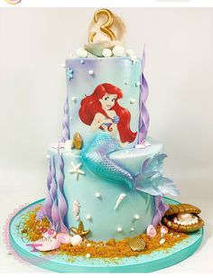 a blue cake with an ariel the mermaid on it's side and under sea shells