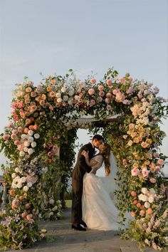 a bride and groom kissing under an arch with flowers