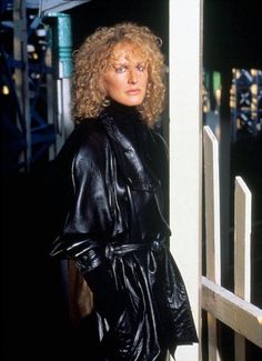 a woman standing next to a white fence wearing a black leather jacket and pants with her hands in her pockets