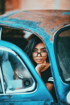 a woman with glasses leaning out the window of an old car looking at the camera