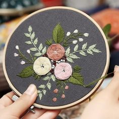 a woman is holding up a hand embroidered flower on a black hoop with green leaves and pink flowers