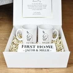 two white coffee mugs in a box with the first home tag on it's side