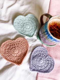 three crocheted heart coasters next to a cup of coffee on a bed