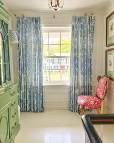 a living room with blue and white curtains on the windowsill, pink chair, and green china cabinet