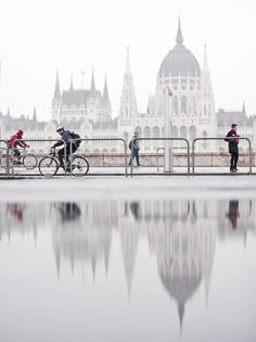 three people riding bikes on a bridge over water in front of a large building with spires