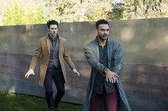 two men dressed in period clothing walking next to a wall and grass area with trees in the background