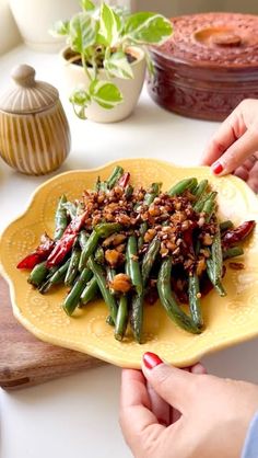 two hands holding a yellow plate with green beans and red peppers on it next to other dishes