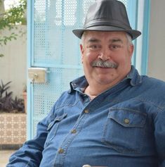 a man with a mustache and a hat sitting in front of a blue wall wearing a denim shirt