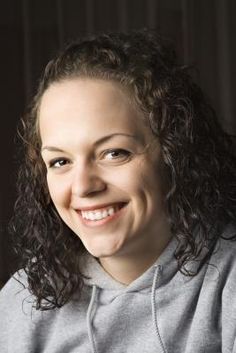 a woman with curly hair smiling at the camera while wearing a gray sweatshirt and silver necklace