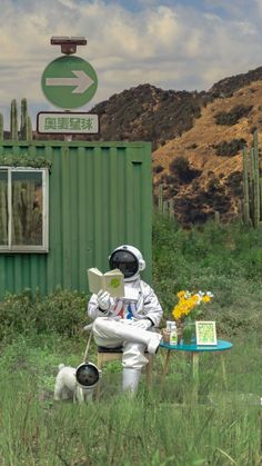 an astronaut sitting on a chair reading a book in front of a green shipping container