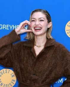 a woman in a brown coat smiling and holding her hand up to her face while standing next to a blue wall