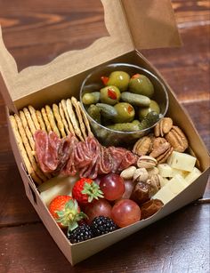 an open box filled with assorted food on top of a wooden table next to a bowl of fruit and crackers