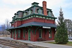 a train station with a red building and green trim on the roof, next to railroad tracks