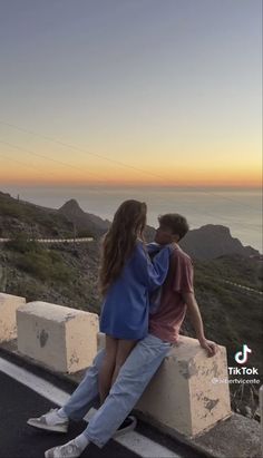 a man and woman sitting on the side of a road looking out at the ocean