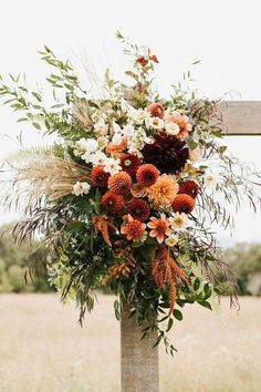 an arrangement of flowers and greenery hangs from a wooden post in the middle of a field