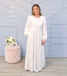 a woman standing in front of a wall wearing a white dress and smiling at the camera