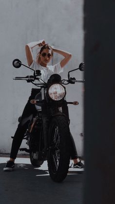 a woman sitting on the back of a motorcycle in front of a white wall and wearing sunglasses