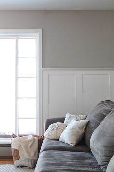 a living room with a gray couch and white paneled wall behind the sofa is a window