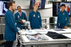 four doctors in scrubs standing around a hospital table with monitors on the wall behind them