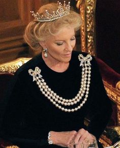 an older woman wearing a tiara and pearls sitting in a chair with her hands together