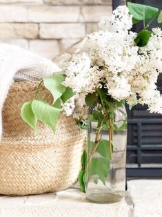 a glass vase with white flowers and green leaves in front of a brick fireplace mantel