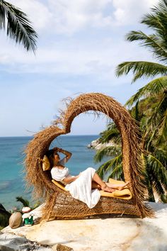 a woman laying on top of a straw covered bench next to the ocean and palm trees