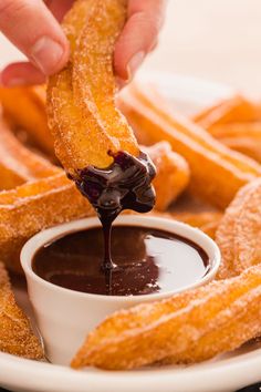 someone dipping chocolate sauce on top of some churros in a white bowl with other churros