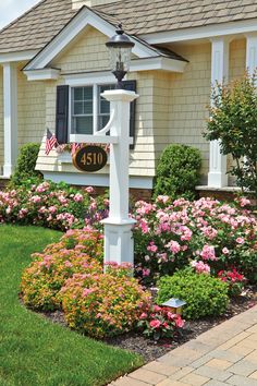 a white mailbox in front of a house with flowers and shrubs around the post