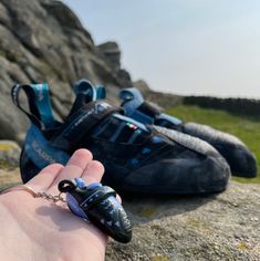 a hand is holding an object in front of two shoes on the ground, with grass and rocks behind them