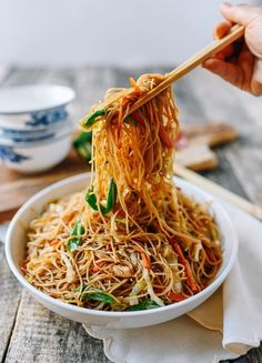 someone holding chopsticks over a bowl of noodles with vegetables and sauce on the side