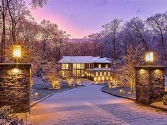 a large house surrounded by trees covered in snow at night with lights on the driveway