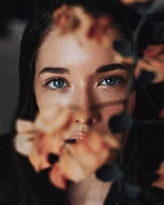 a woman with blue eyes looking at the camera through some leaves and flowers in front of her face