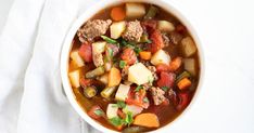 a white bowl filled with meat and vegetable soup on top of a white cloth next to a spoon