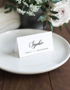 a place card sitting on top of a white plate next to a vase with flowers