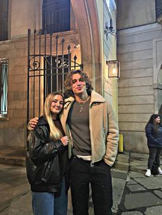 a man and woman standing in front of a building at night with their arms around each other