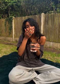 a woman sitting on the ground holding a glass of water in her hand and covering her mouth with both hands