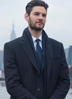 a man wearing a suit and tie standing in front of a body of water with a cityscape in the background