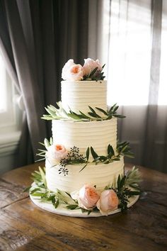 a three tiered cake with flowers and greenery on the top is sitting on a wooden table