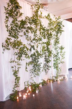 candles are lit in front of a white backdrop with greenery on it and hanging from the ceiling