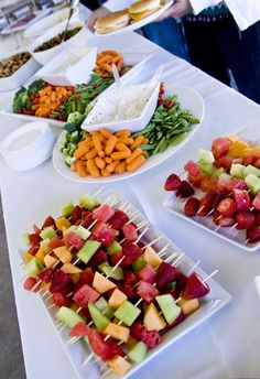 various fruits and vegetables are arranged on trays at a buffet table with people in the background