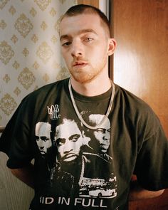 a man in a black t - shirt is looking at the camera while standing next to a door