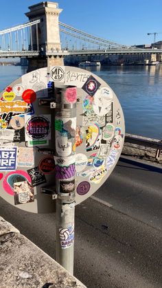 a street sign covered in stickers next to a body of water with a bridge in the background