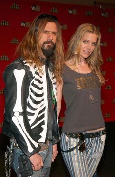 a man and woman standing next to each other in front of a red wall with a skeleton on it